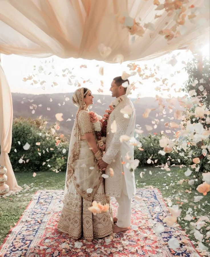 Bride & groom in traditional attire under a canopy with flower petals.